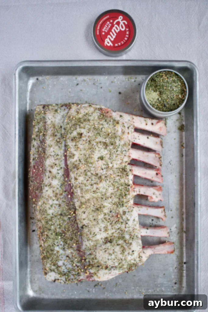 Rack of Lamb seasoned generously with fragrant herbs and spices, resting on a cutting board to reach room temperature before cooking.