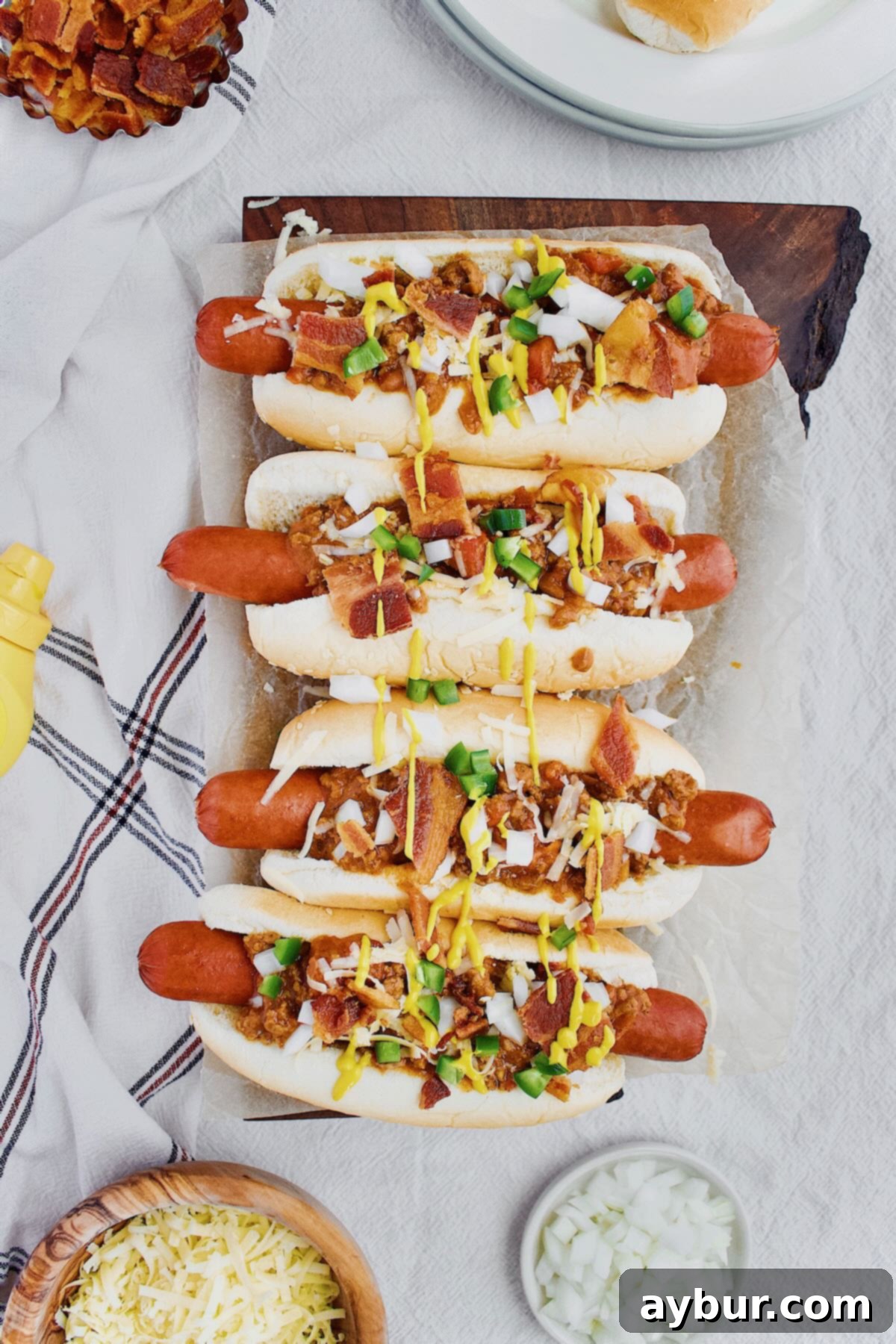 A vibrant spread of chili dogs generously topped with cheddar cheese, finely diced onions, spicy jalapeños, classic yellow mustard, and a hearty dollop of homemade Hot Dog Sauce.