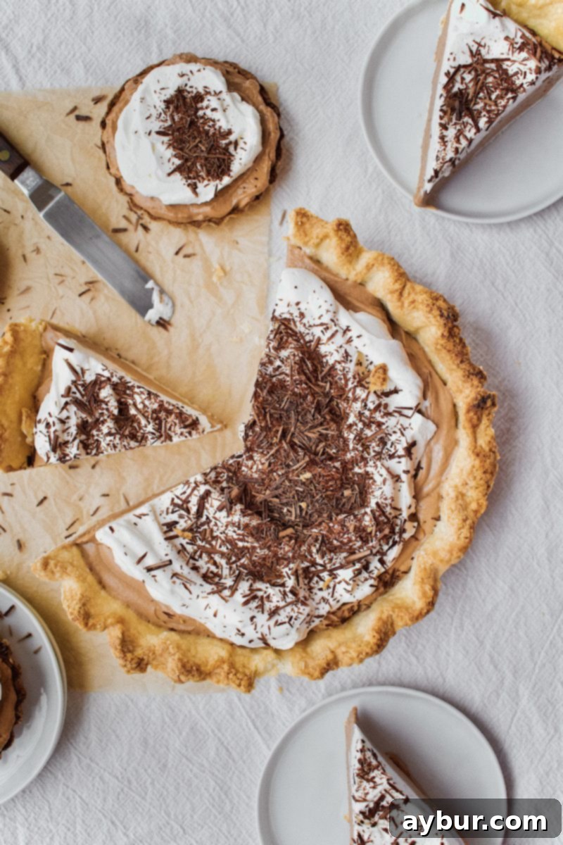 A perfectly sliced Joanna Gaines French Silk Pie, rich chocolate filling topped with whipped cream, surrounded by miniature tartlets on a rustic wooden surface.