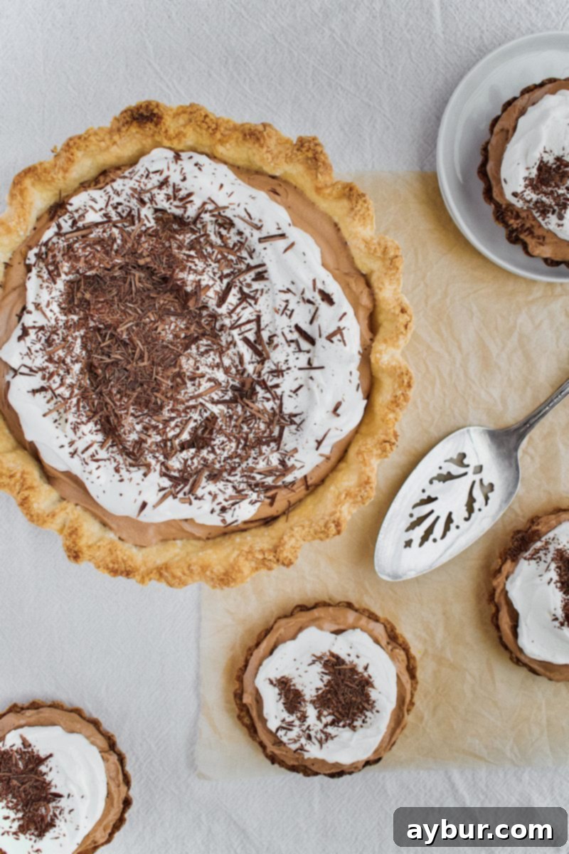 Close-up of a French Silk Pie slice, showing the layers of crisp crust, rich chocolate filling, and fluffy whipped cream, next to mini chocolate tartlets.