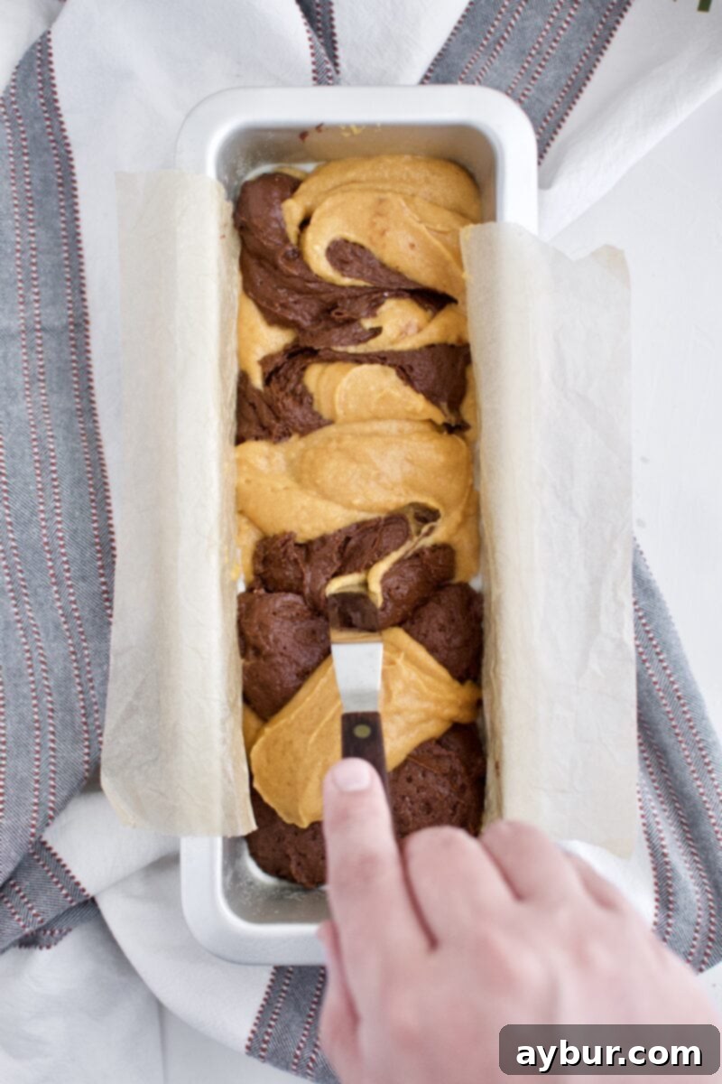 A knife gently swirling pumpkin and chocolate batters together in a loaf pan to create a marbled effect