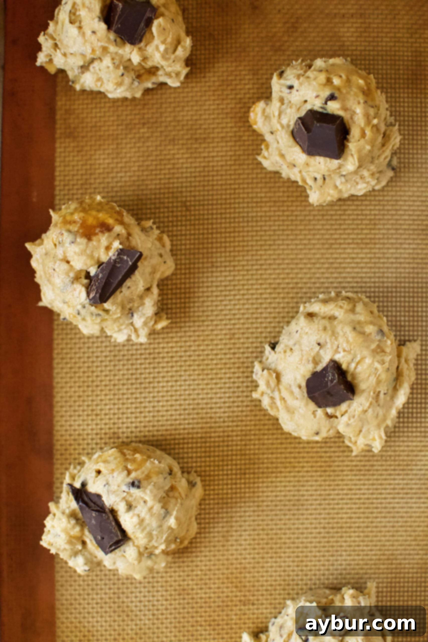 Portioned cookie dough balls on a parchment-lined baking tray, each topped with a single large chocolate chunk, ready for baking.