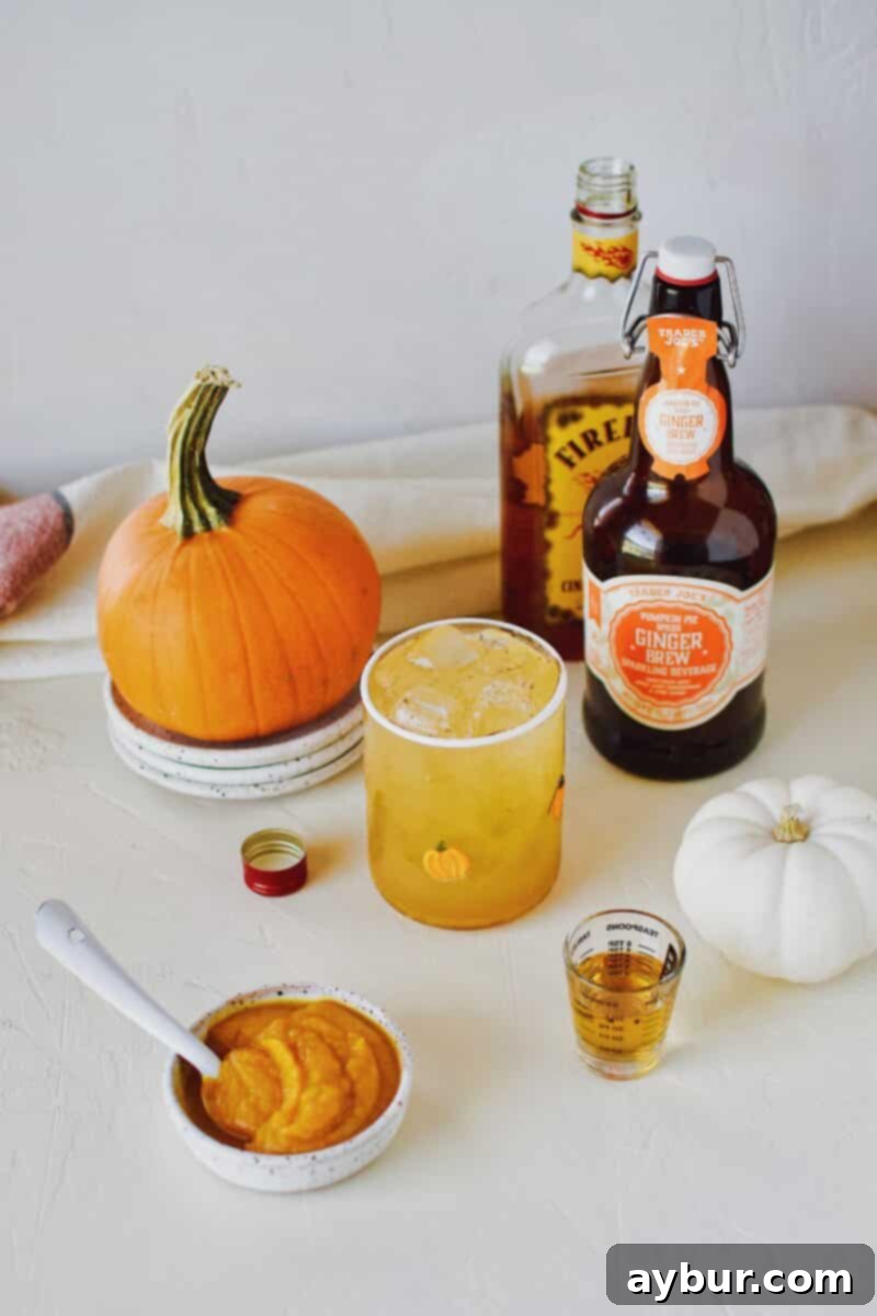 Close-up of a glass of spiked sparkling pumpkin juice garnished with a cinnamon stick, featuring bottles of pumpkin spice ginger beer and cinnamon whisky in the background.