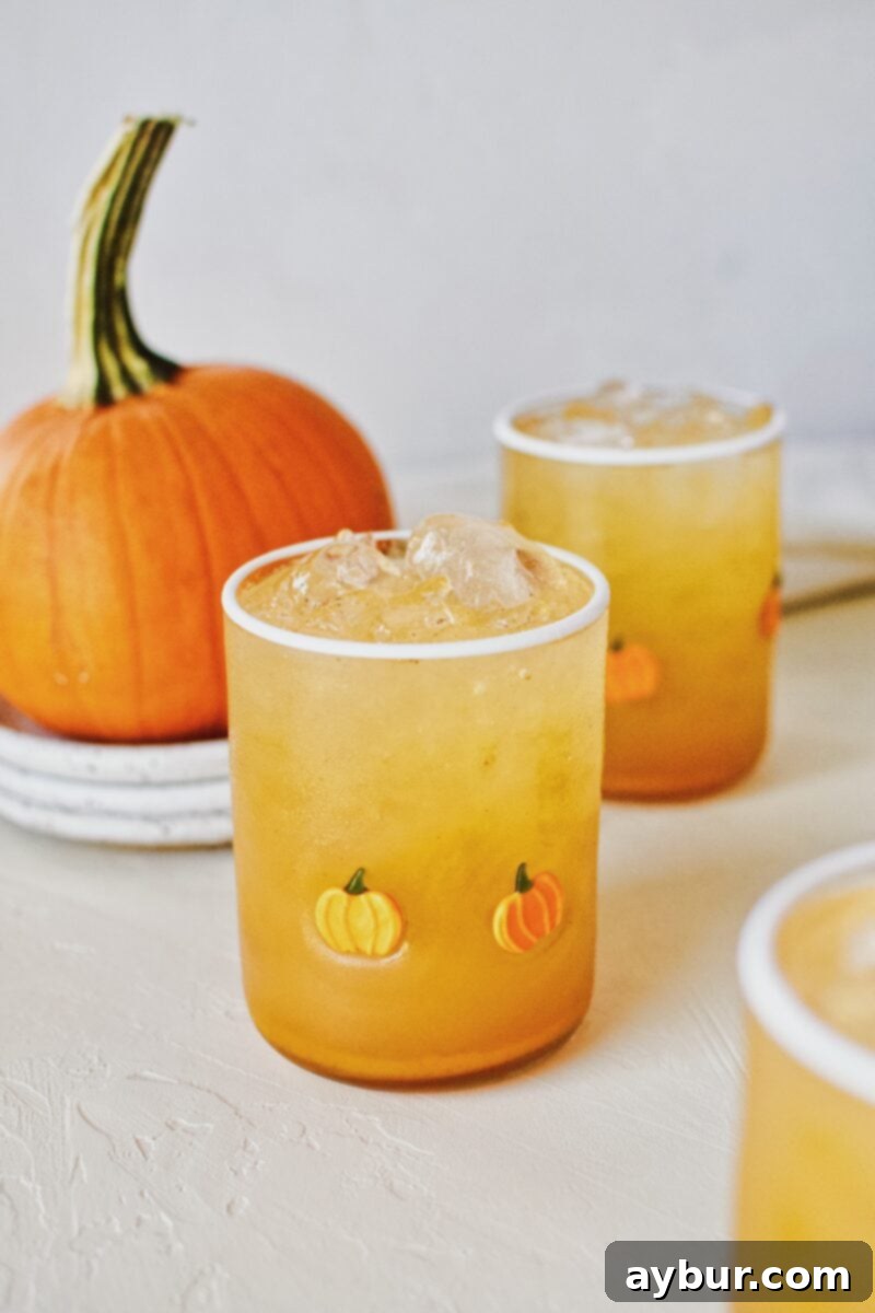 A glass of homemade pumpkin juice in a unique pumpkin-shaped glass, positioned next to a small, ornamental pie pumpkin.