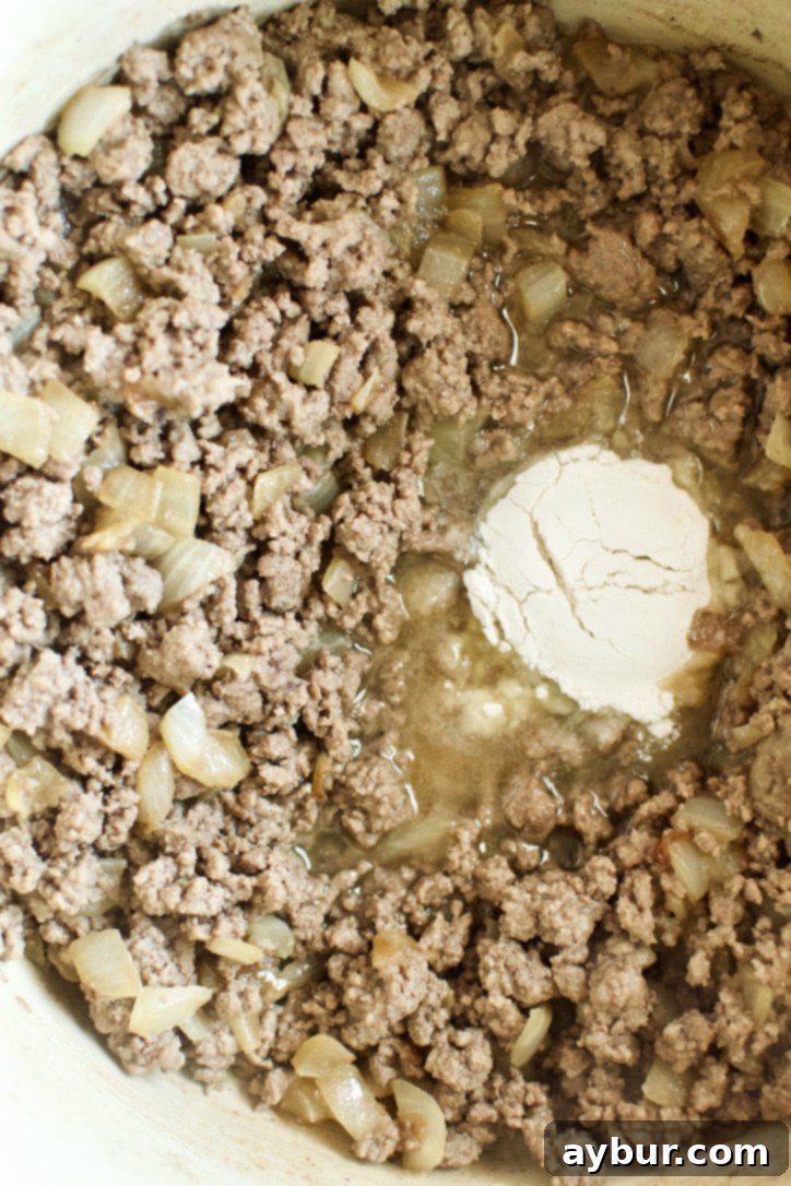 Ground beef cooking with sautéed onions in a pot, then flour is sprinkled in to thicken the mixture.