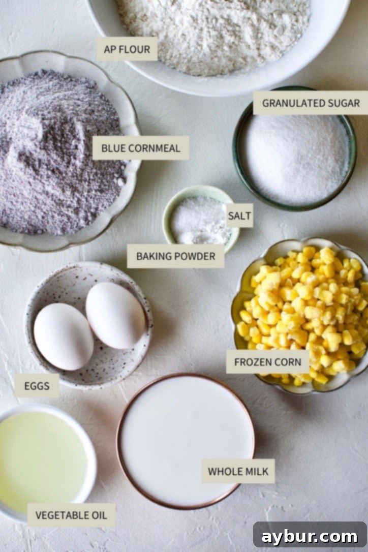 All essential ingredients for making blue cornbread laid out on a kitchen counter, including milk, oil, eggs, blue cornmeal, flour, sugar, and corn kernels.