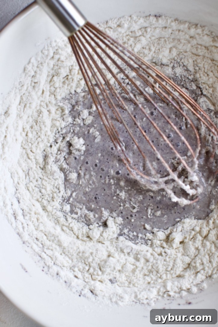 Dry ingredients for the blue cornbread, including cornmeal and flour, being carefully whisked into the wet mixture in a single bowl.