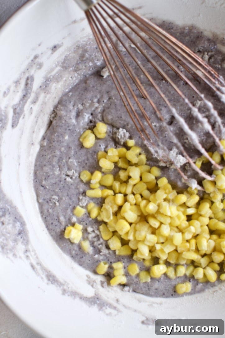 Frozen corn kernels being gently folded into the blue cornbread batter using a spatula.