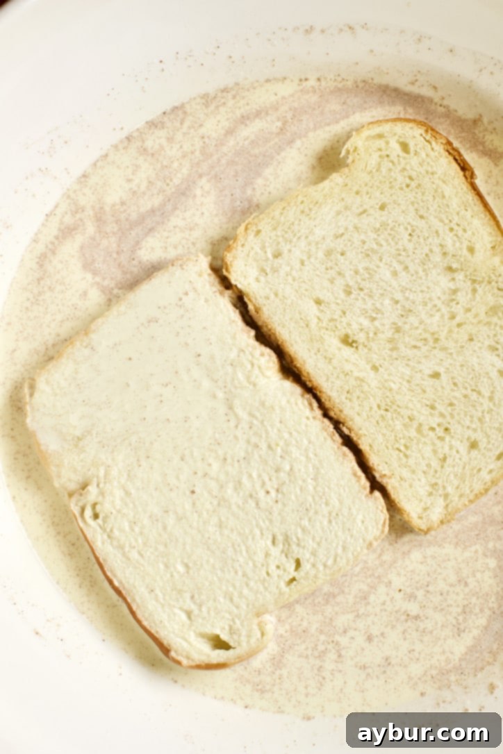 A slice of brioche bread being carefully dipped and submerged into the rich, frothy custard mixture in a shallow dish, ensuring even saturation.