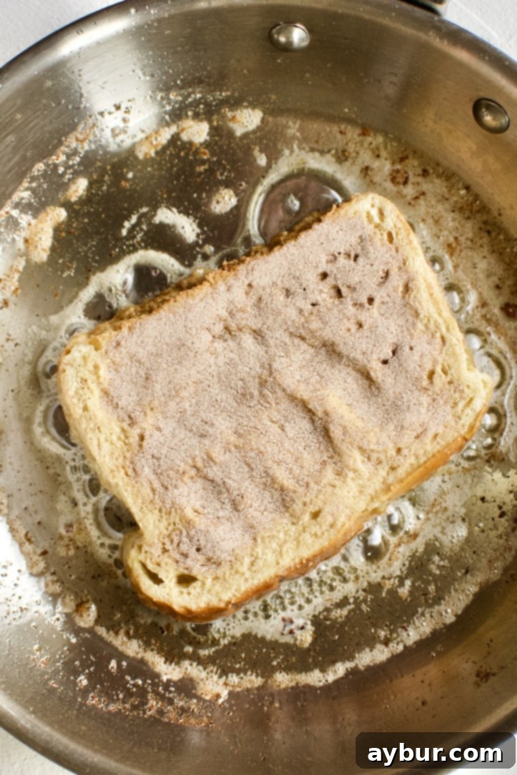 Two slices of French Toast cooking in a skillet, with the first side turning golden brown and the second side lightly dusted with cinnamon sugar, preparing for the flip.