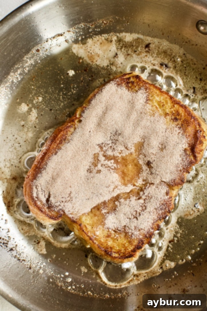 One slice of French Toast flipped in the pan, revealing a perfectly golden and caramelized underside, with the top side now dusted with cinnamon sugar, continuing to cook.