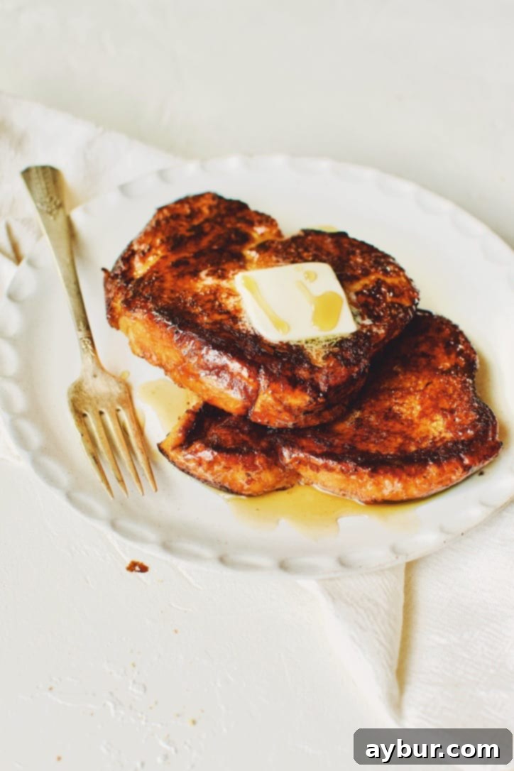 A close-up of a stack of caramelized Custard French Toast slices, elegantly topped with a square of melting butter and glistening with maple syrup.