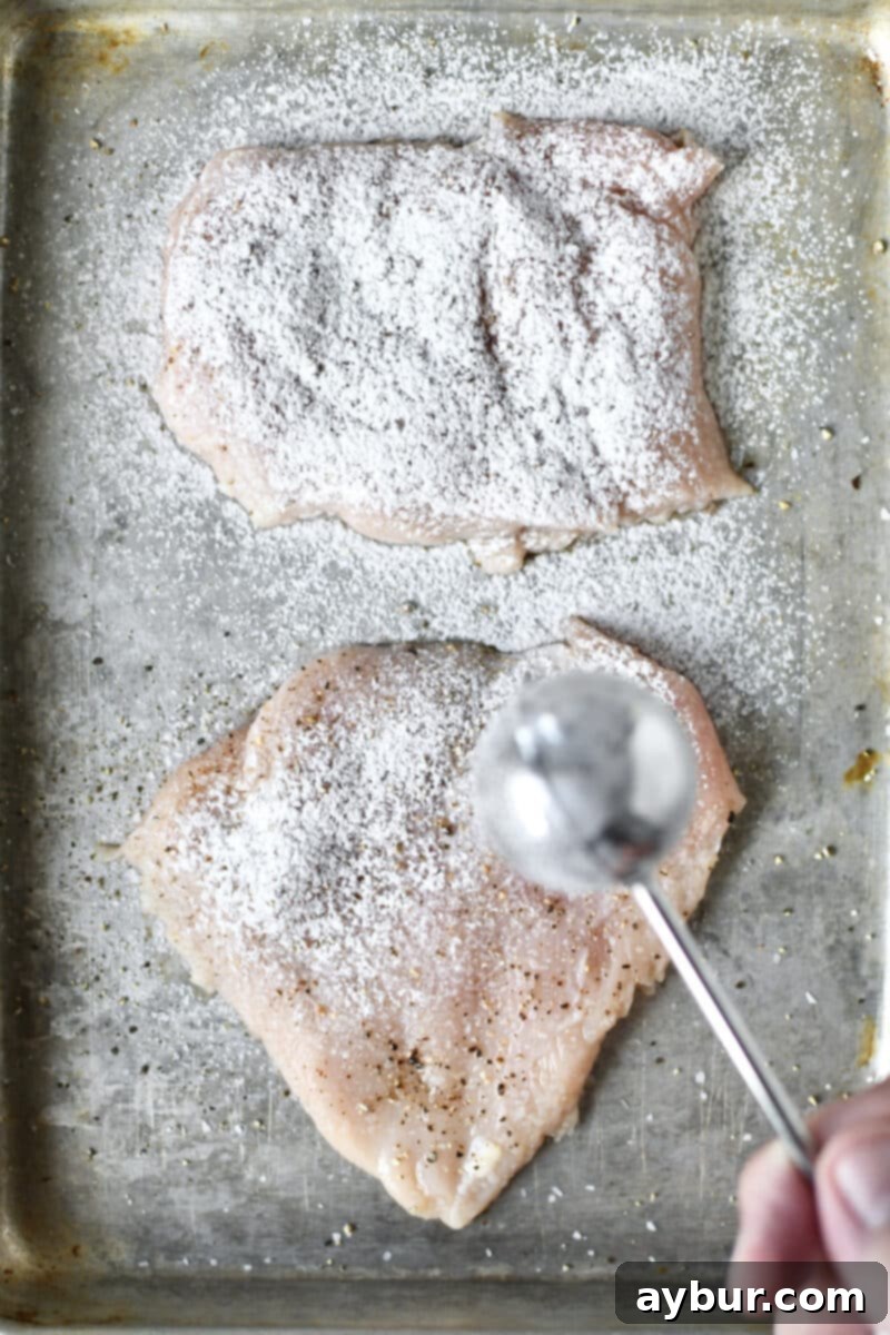 Dusting a seasoned, tenderized chicken breast with flour using a dusting wand for even coating.