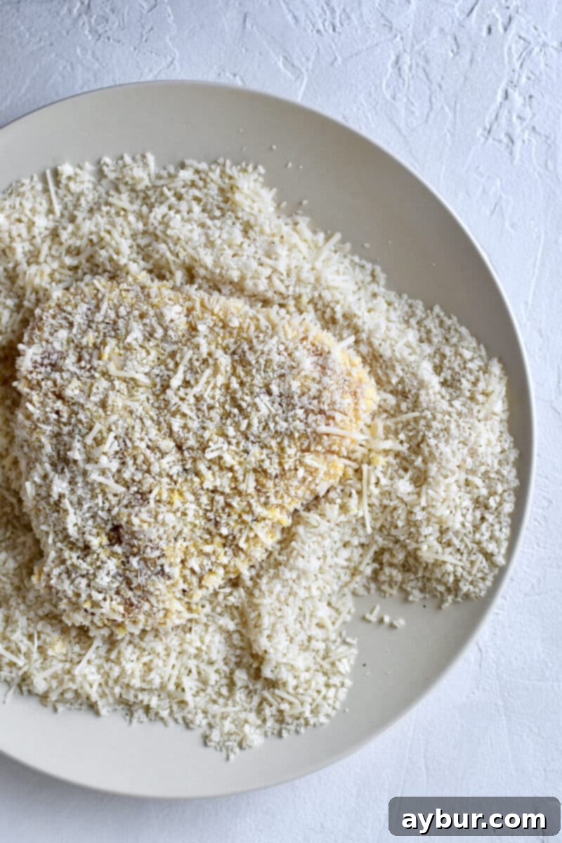 Chicken breast being coated with parmesan and breadcrumbs, pressing firmly for a full crust.