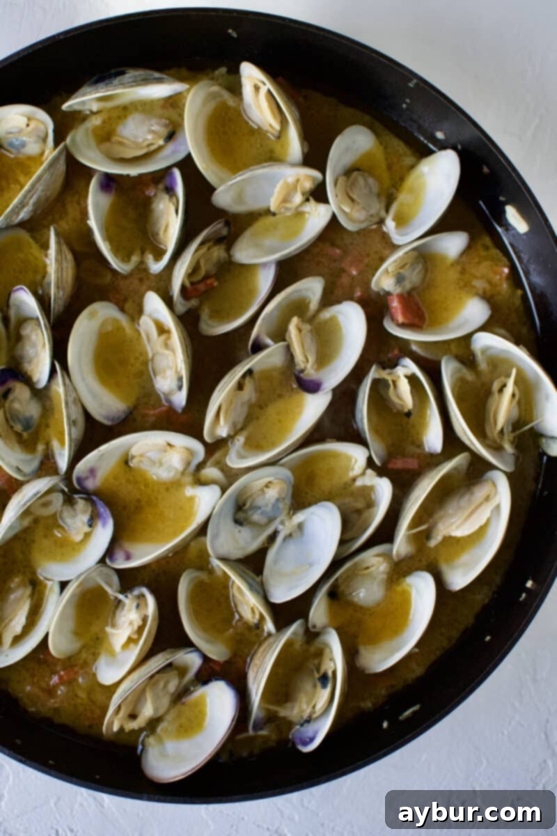 Linguine with Clams 11 An open pan revealing a vibrant mixture of opened clams, cooked garlic, onions, and chorizo, all simmering in a delicious white wine sauce, indicating the clams are perfectly steamed and ready for pasta.