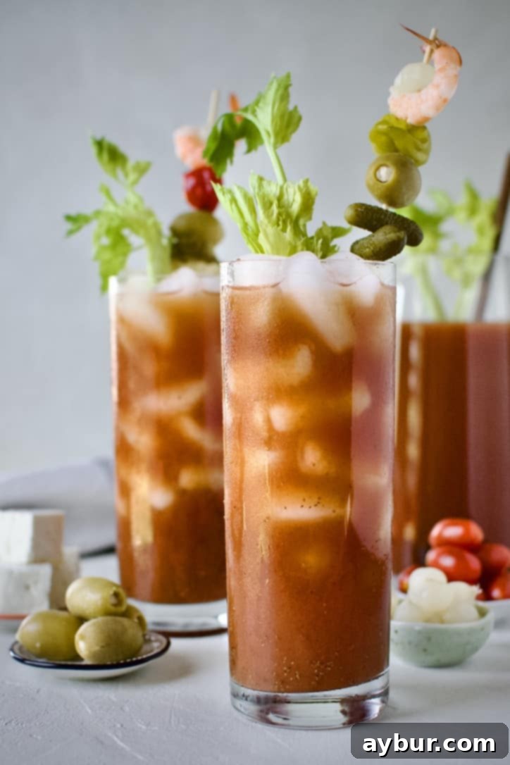 Two perfectly garnished Bloody Mary cocktails, brimming with savory toppings like pickles, olives, peppers, and shrimp, set against a blurred background of a pitcher filled with Bloody Mary mix.