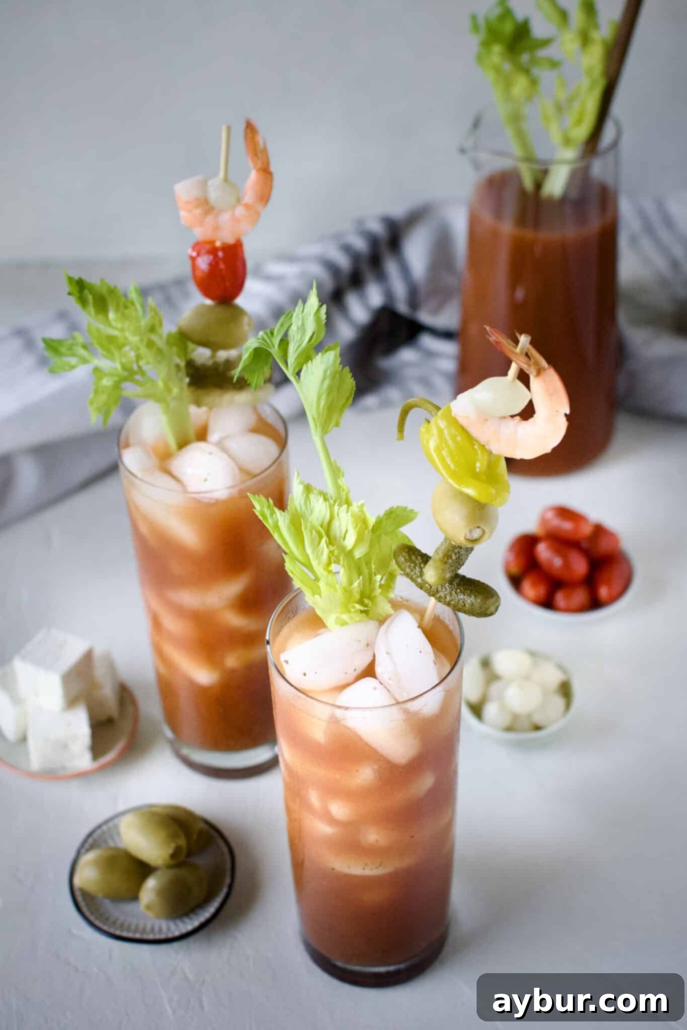 A vibrant pitcher of freshly mixed Bloody Mary cocktail stands ready, surrounded by glasses generously adorned with an array of appealing garnishes like crispy bacon, plump shrimp, various pickles, olives, and colorful peppers.