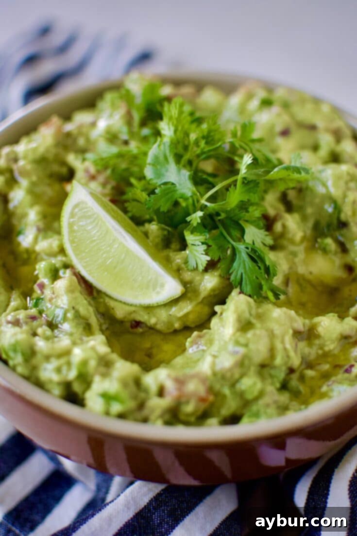 Mexican Guacamole Recipe in a bowl topped with a lime wedge, baby cilantro sprigs and olive oil.