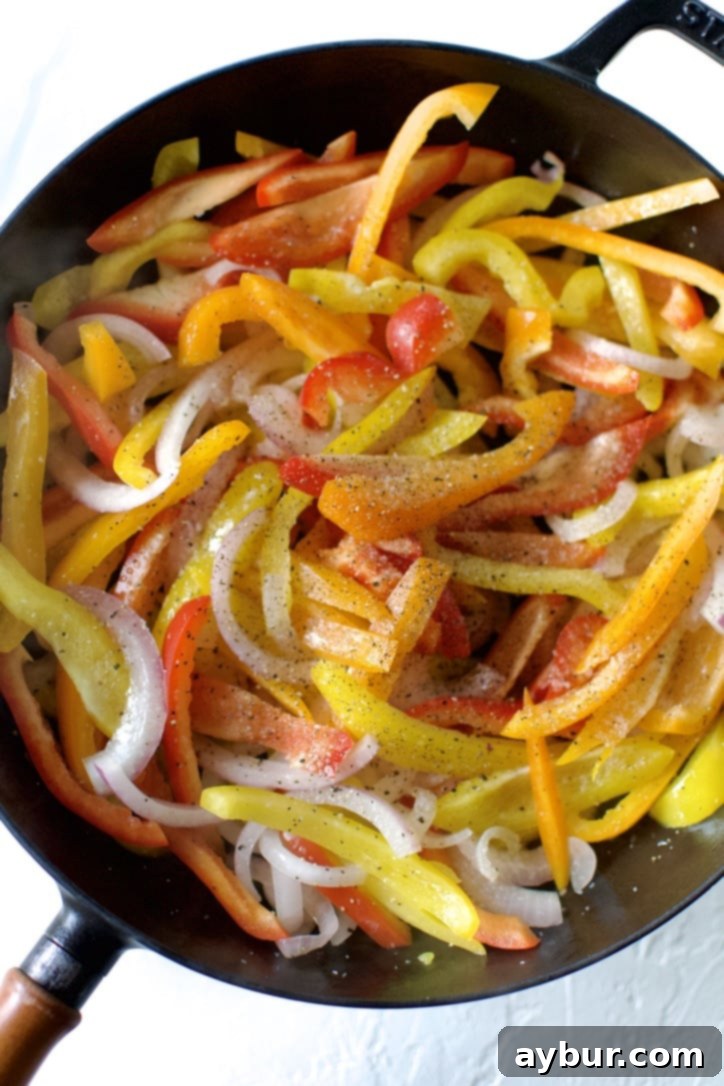 Sliced red onions and colorful bell peppers being added to a hot skillet with olive oil, beginning to sauté and soften.