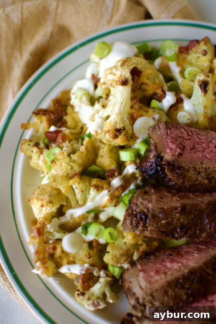 Loaded Baked Cauliflower served on a white plate alongside a perfectly grilled steak.