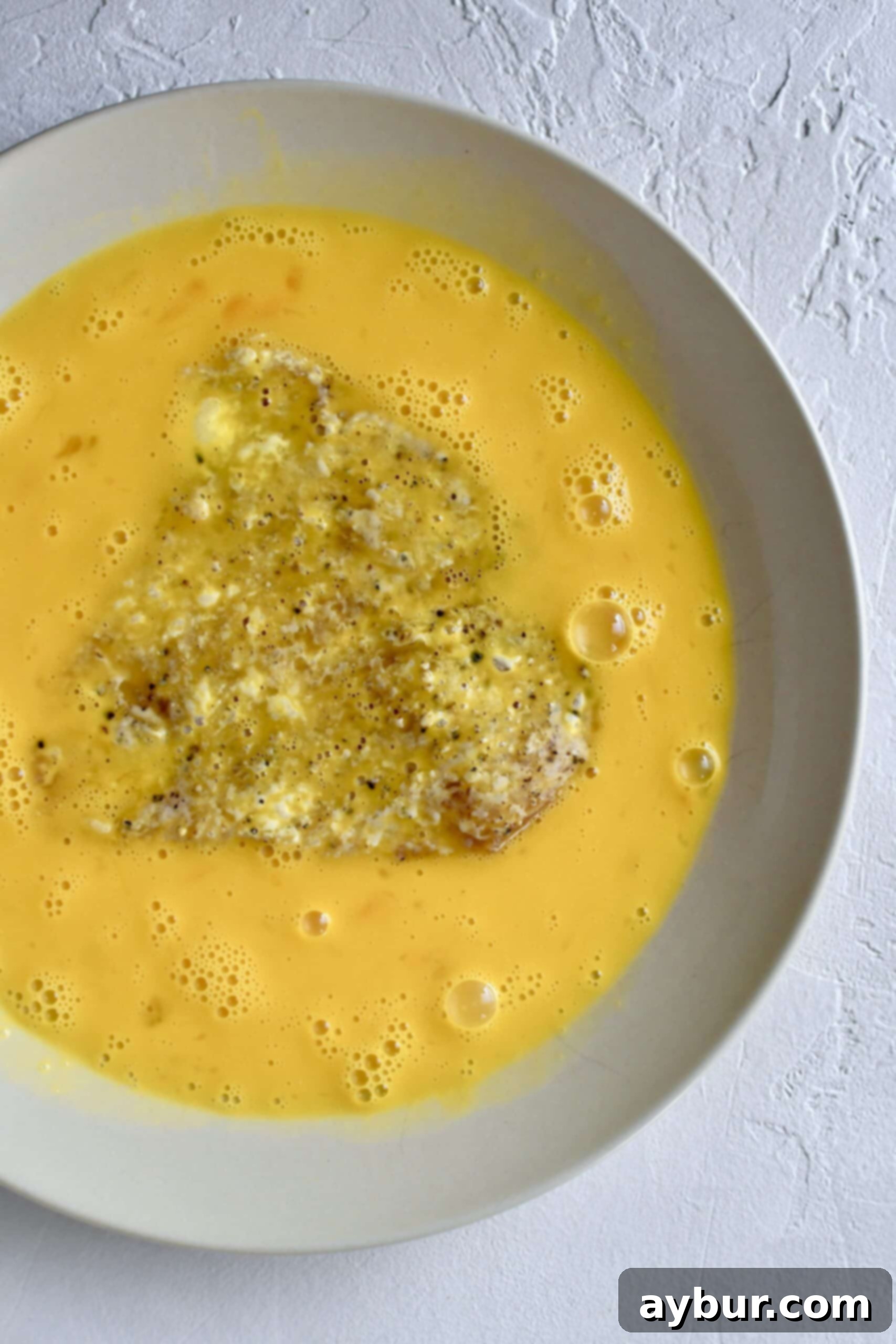 Chicken breast being dredged in beaten eggs as part of the breading process.