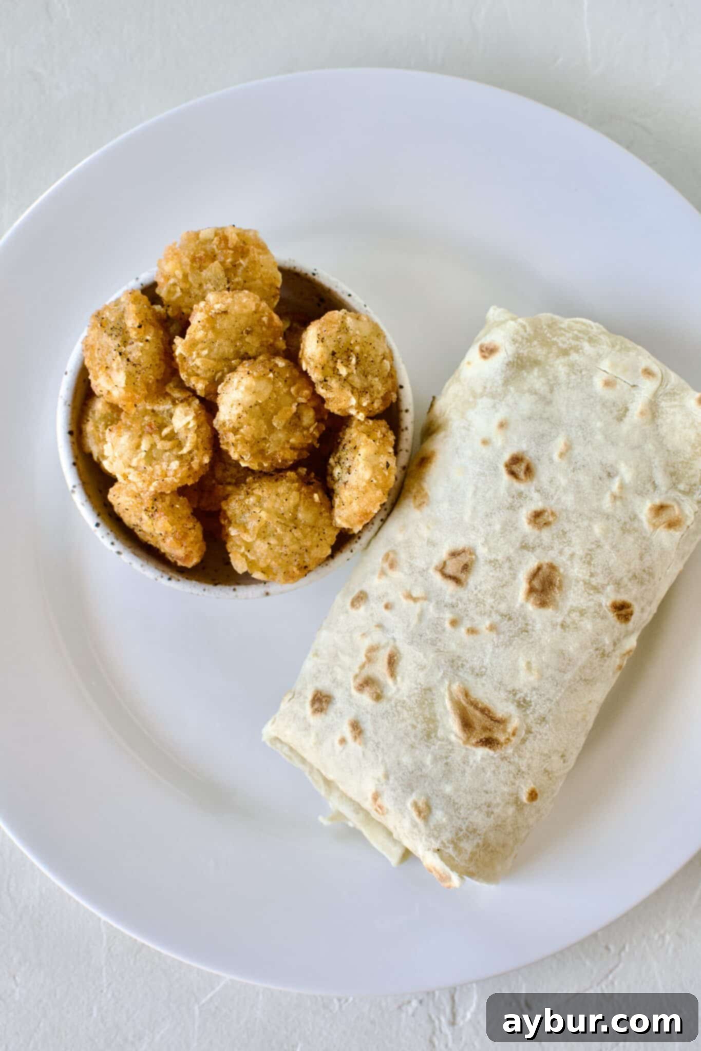 Homemade Breakfast Burrito all rolled up on a plate next to a pile of extra hash browns.
