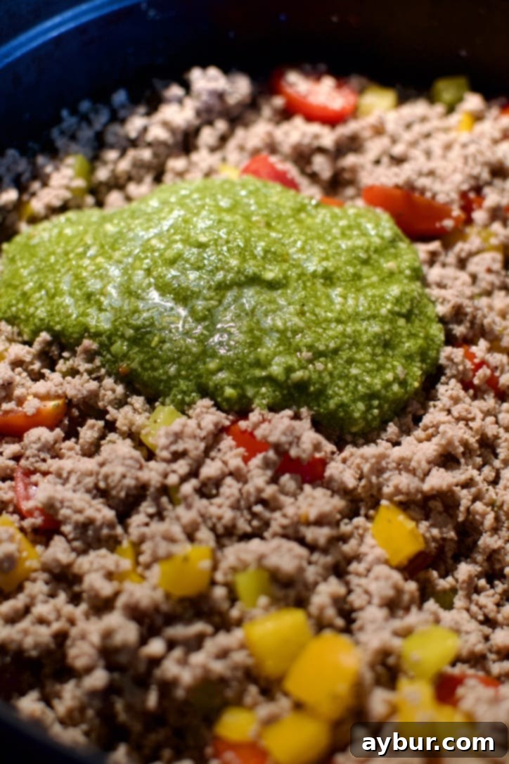 Fresh basil pesto being stirred into the cooked ground turkey and vegetable mixture in a large pot.
