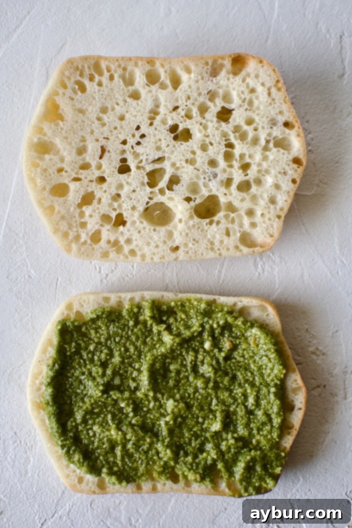 Step 1: Preparing the ciabatta roll. A sliced ciabatta roll with a generous spread of vibrant green pesto sauce on the bottom half, ready for further fillings.