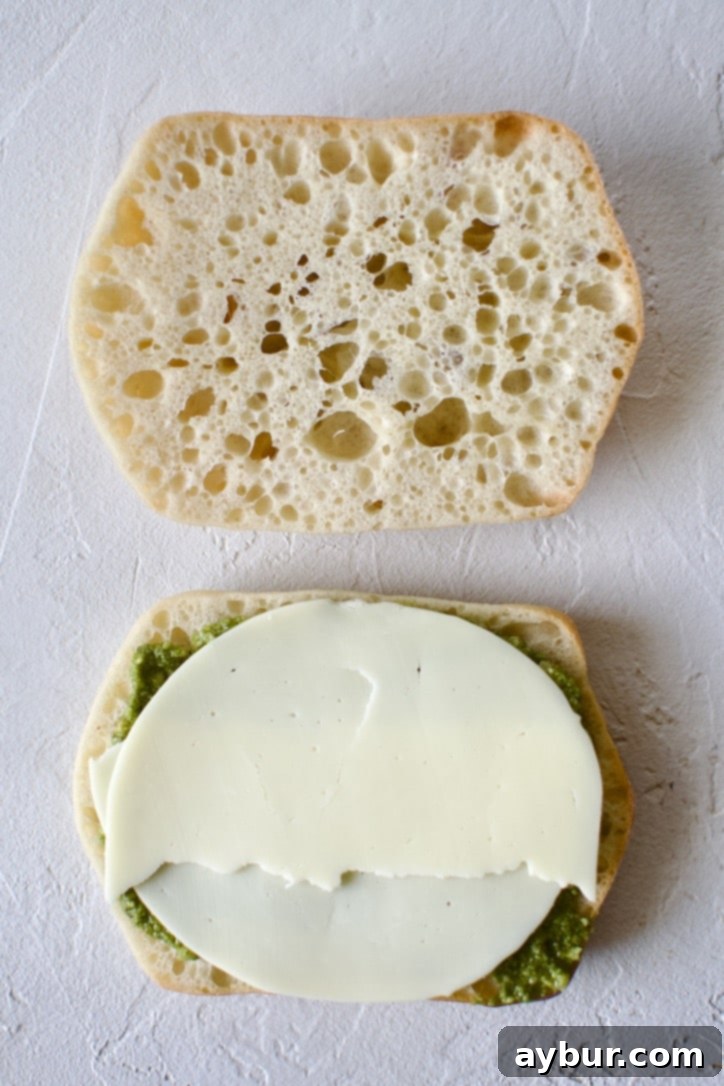 Step 2: Adding the first layer of cheese. A ciabatta roll with pesto sauce, now topped with several slices of provolone cheese, forming a foundational layer.