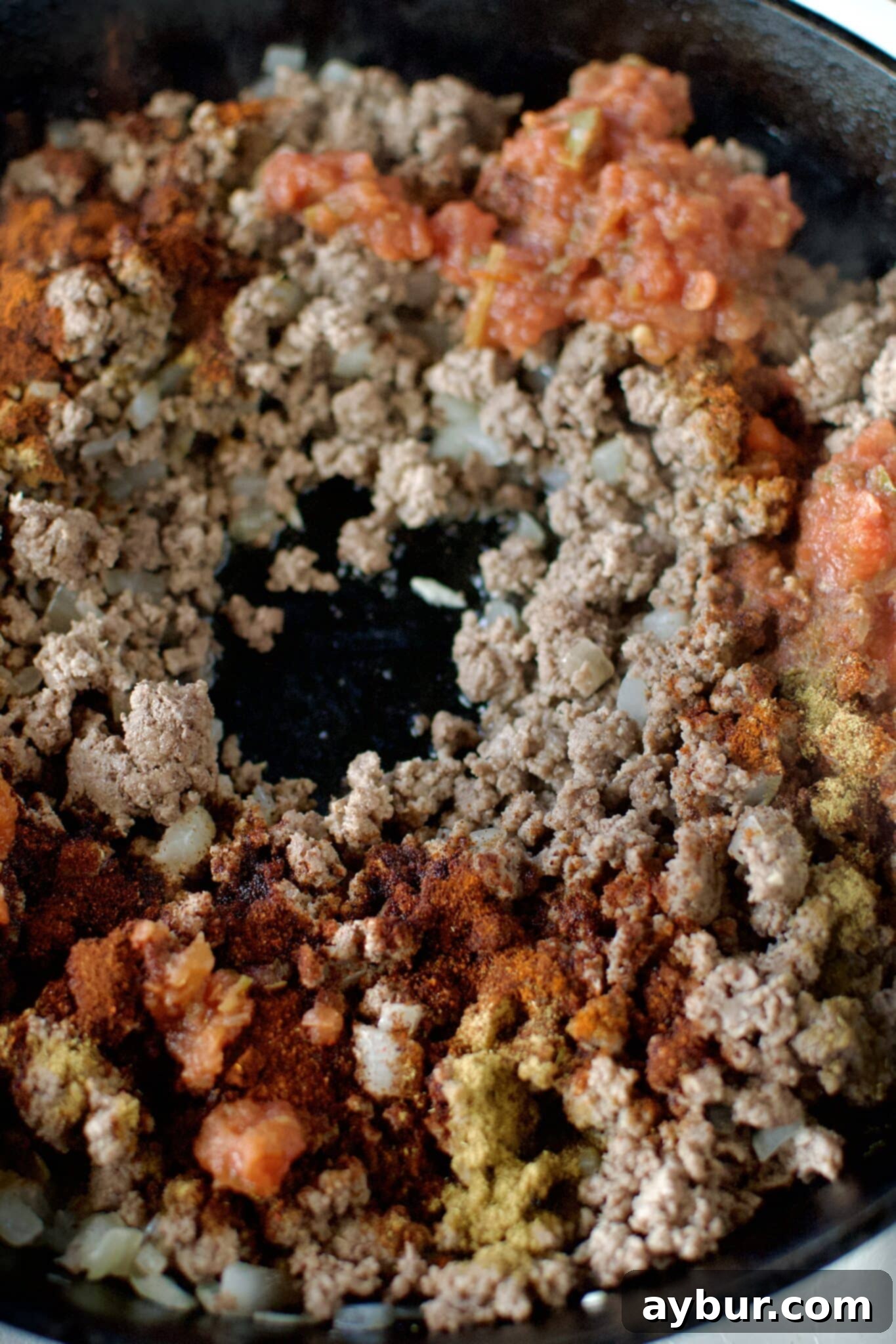 Cooked ground beef in a cast iron skillet, with a spoon ready to add the spices.