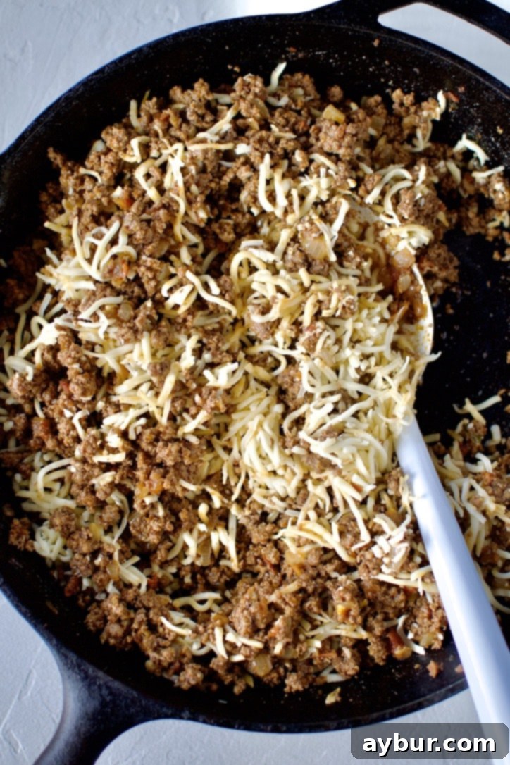Shredded Chihuahua cheese being stirred into the cooked and seasoned ground beef filling in a cast iron skillet.