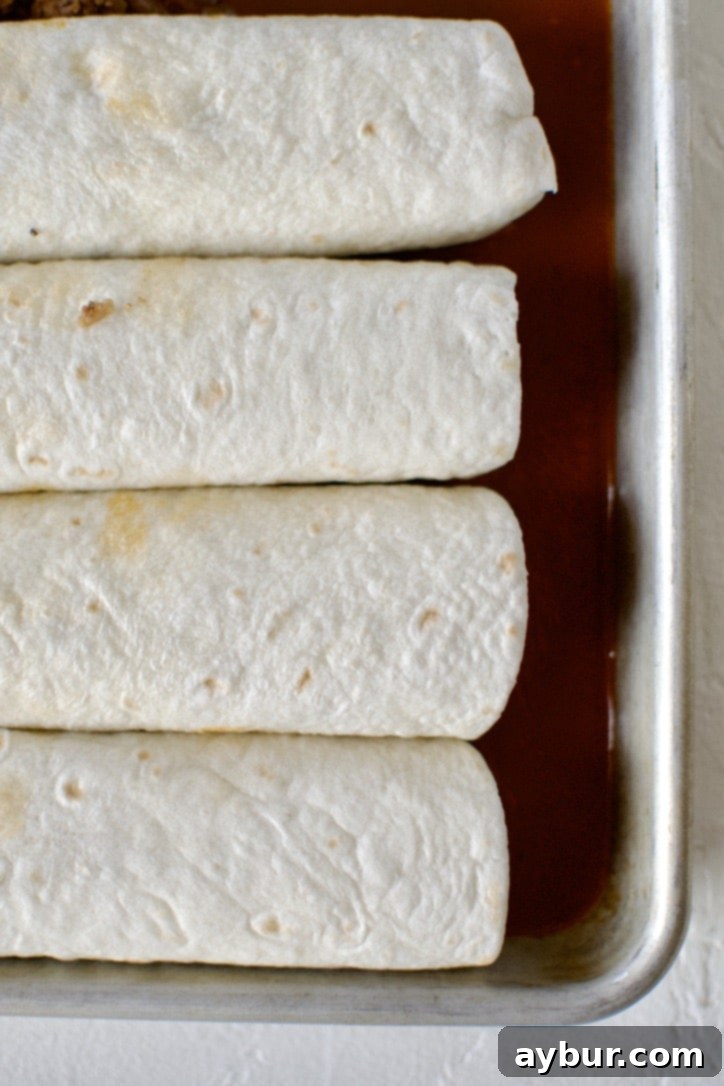 Neatly arranged filled enchiladas in a baking pan, resting on a layer of red enchilada sauce.