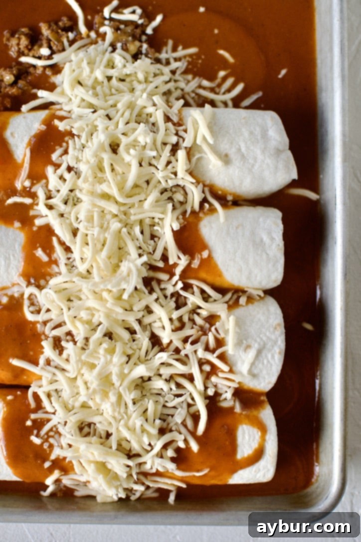 Rolled enchiladas in a baking pan, generously topped with red enchilada sauce and an ample layer of shredded cheese, ready for baking.