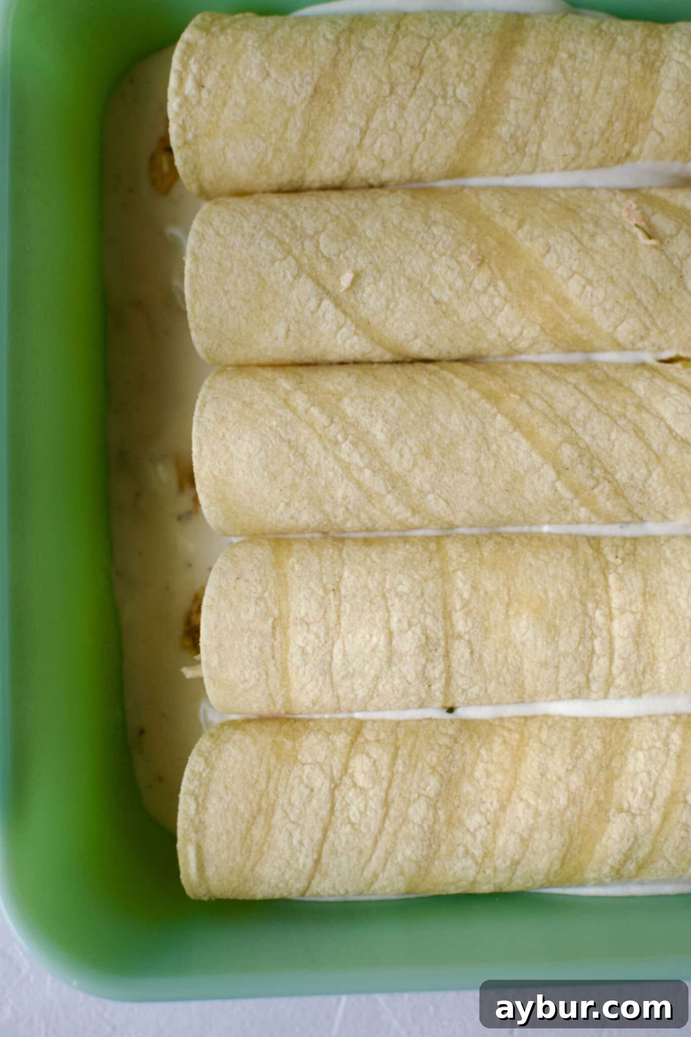 Chicken filled tortillas rolled up and placed in a baking dish that has been lined with sour cream enchilada sauce.