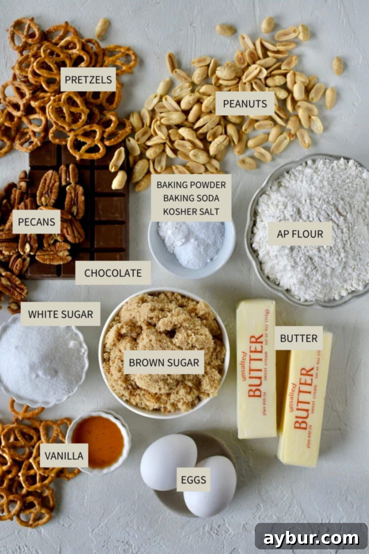 Various ingredients for kitchen sink cookies laid out on a wooden surface, including chocolate, nuts, pretzels, butter, and flour.