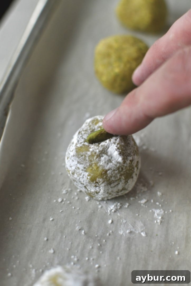 A hand pressing a pistachio halve onto the top of a portioned cookie dough ball.