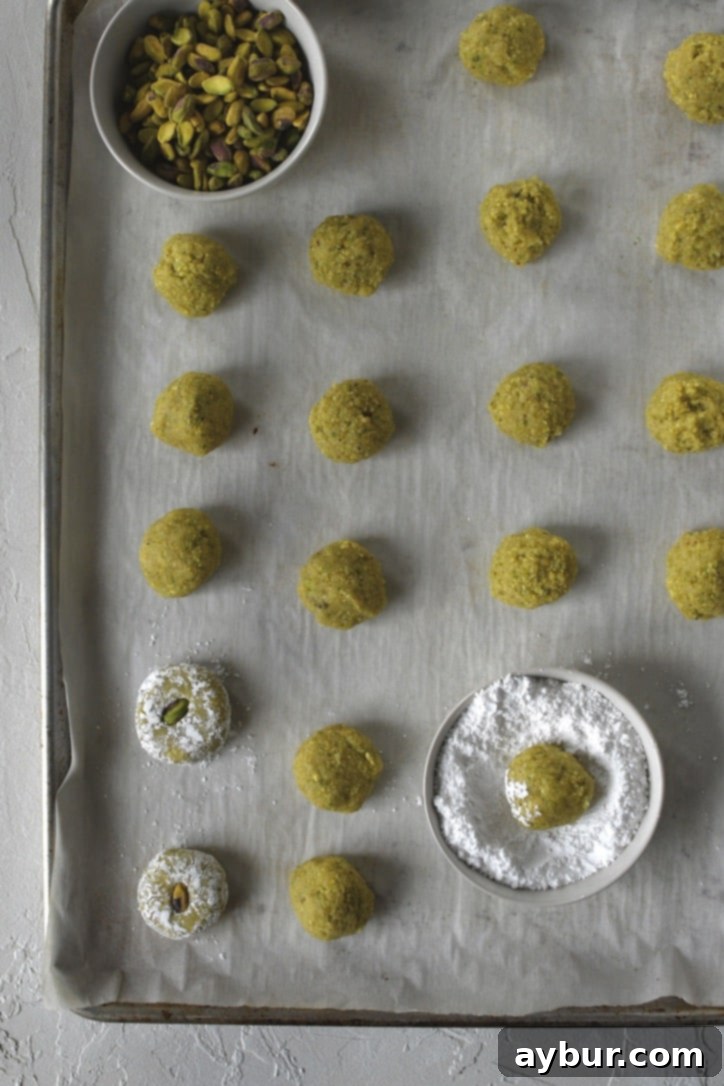 Unbaked Pistachio Amaretti Cookies coated in powdered sugar and topped with pistachio halves, arranged on a baking sheet.