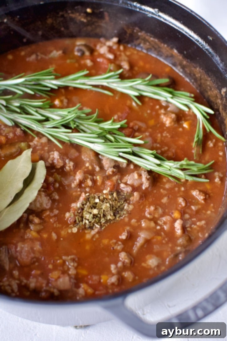 Raw San Marzano tomatoes are being added to the pot of browned sausage and vegetables, with whole bay leaves, fresh rosemary sprigs, and red pepper flakes joining the mixture for simmering.