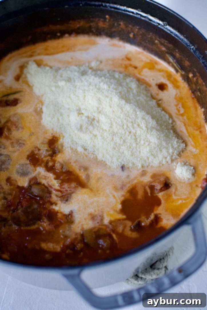 A hand adding a swirl of rich heavy cream and a sprinkle of grated Parmesan cheese into the simmering Sausage Bolognese sauce in a large pot, signifying the final luxurious touches.