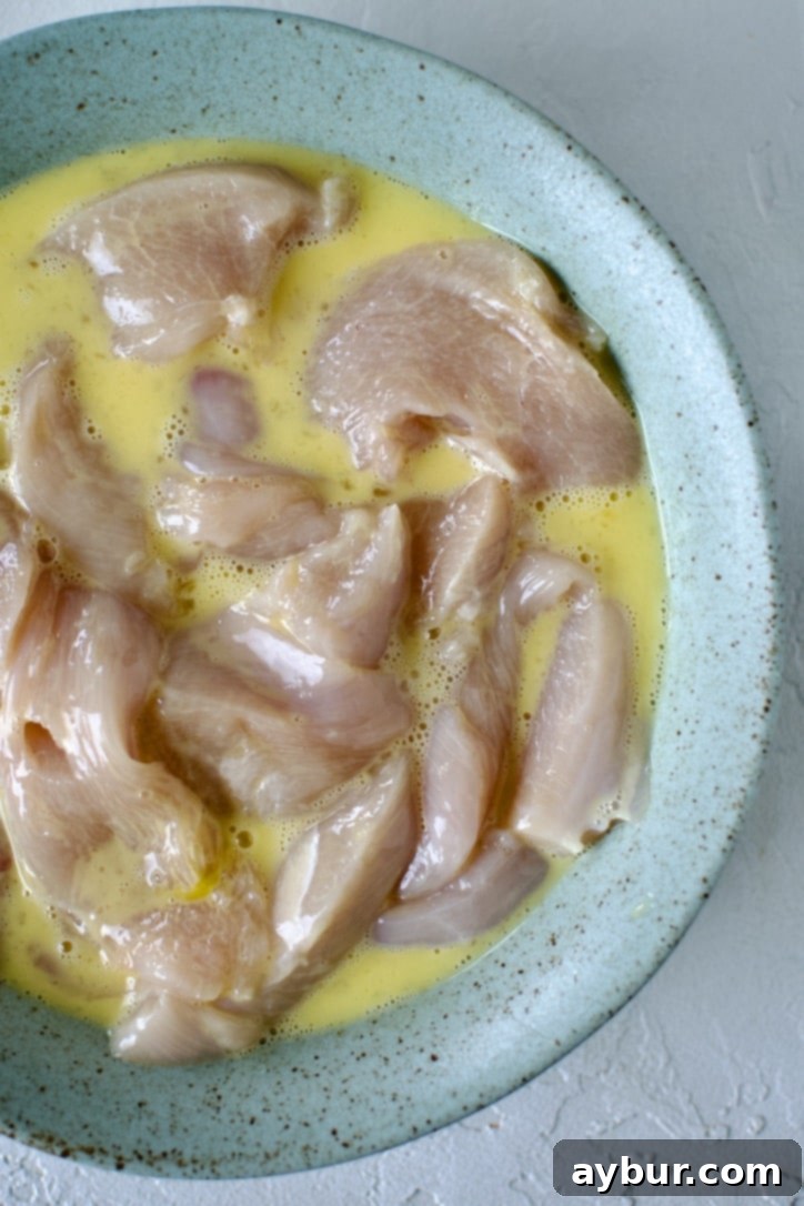 Chicken tenders being dipped into beaten eggs and milk in a shallow bowl as part of the breading process.