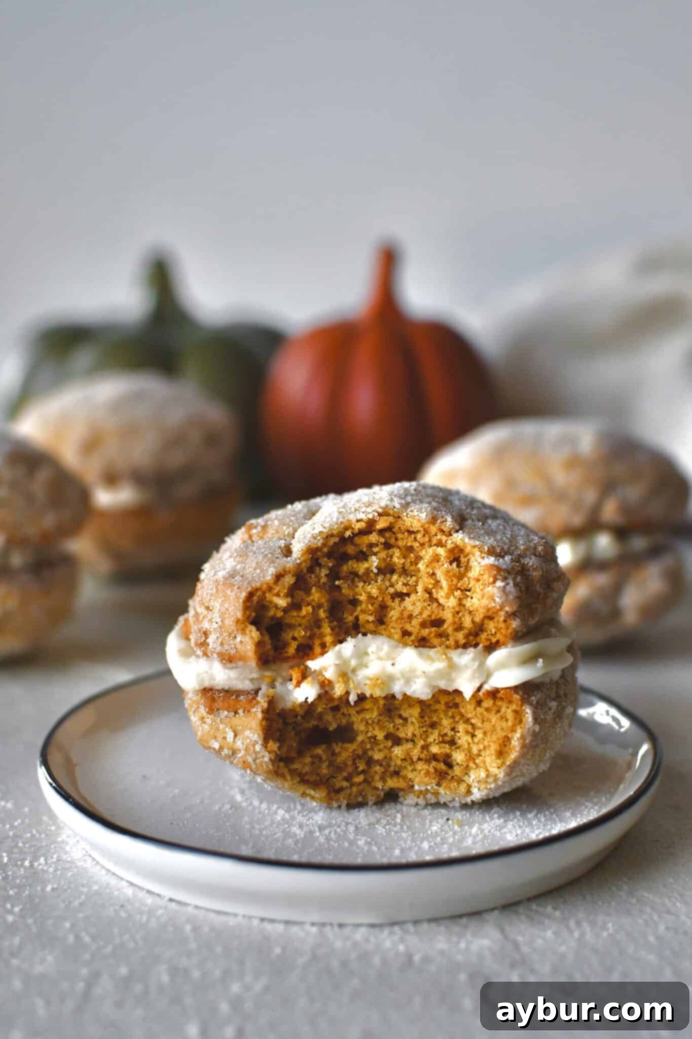 A Pumpkin Whoopie Pie with a bite removed, resting on a plate with more untouched whoopie pies in the background, adorned with fall decor.