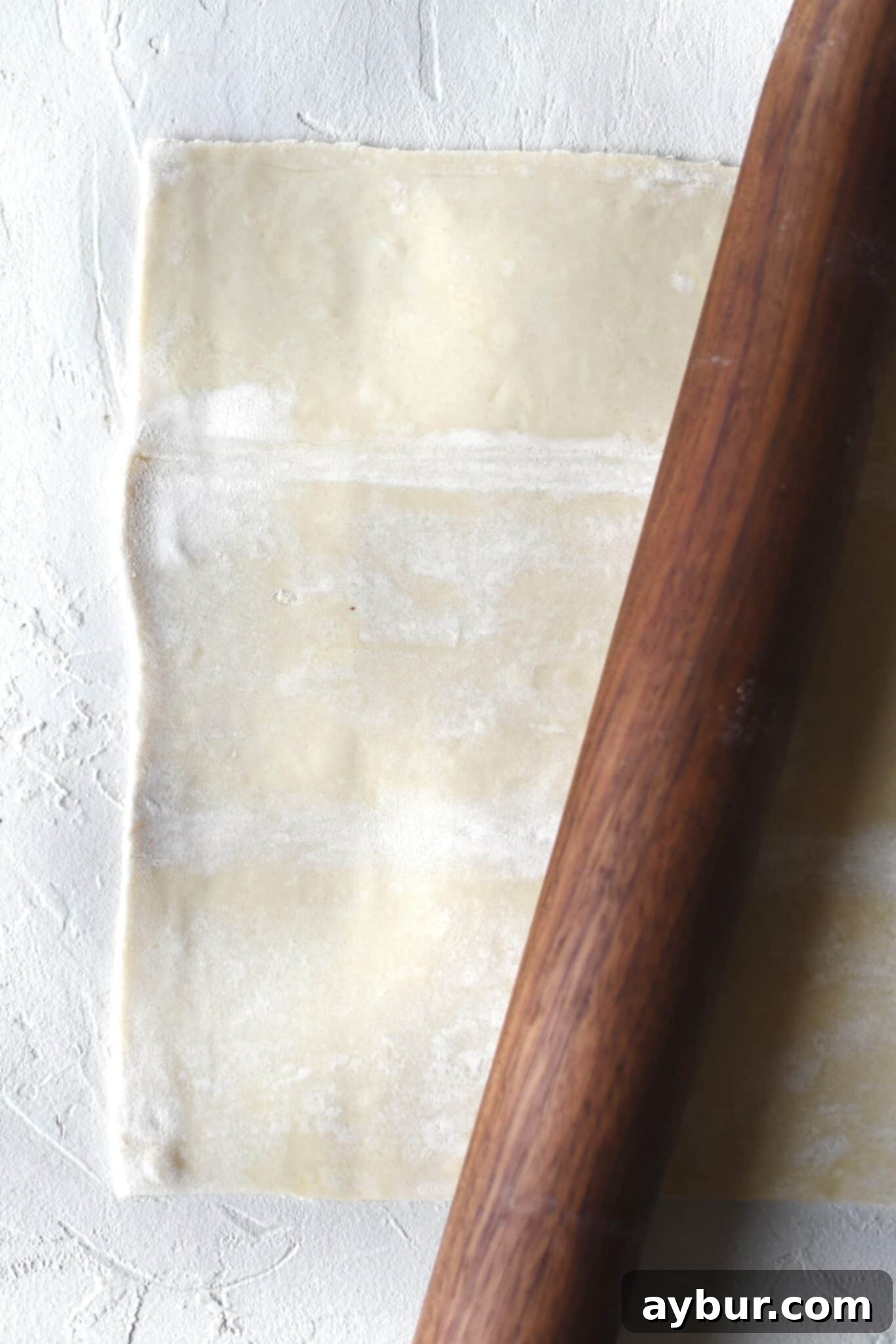 Gently rolling out a sheet of puff pastry on a lightly floured surface to refine its shape and flatten it slightly before cutting.