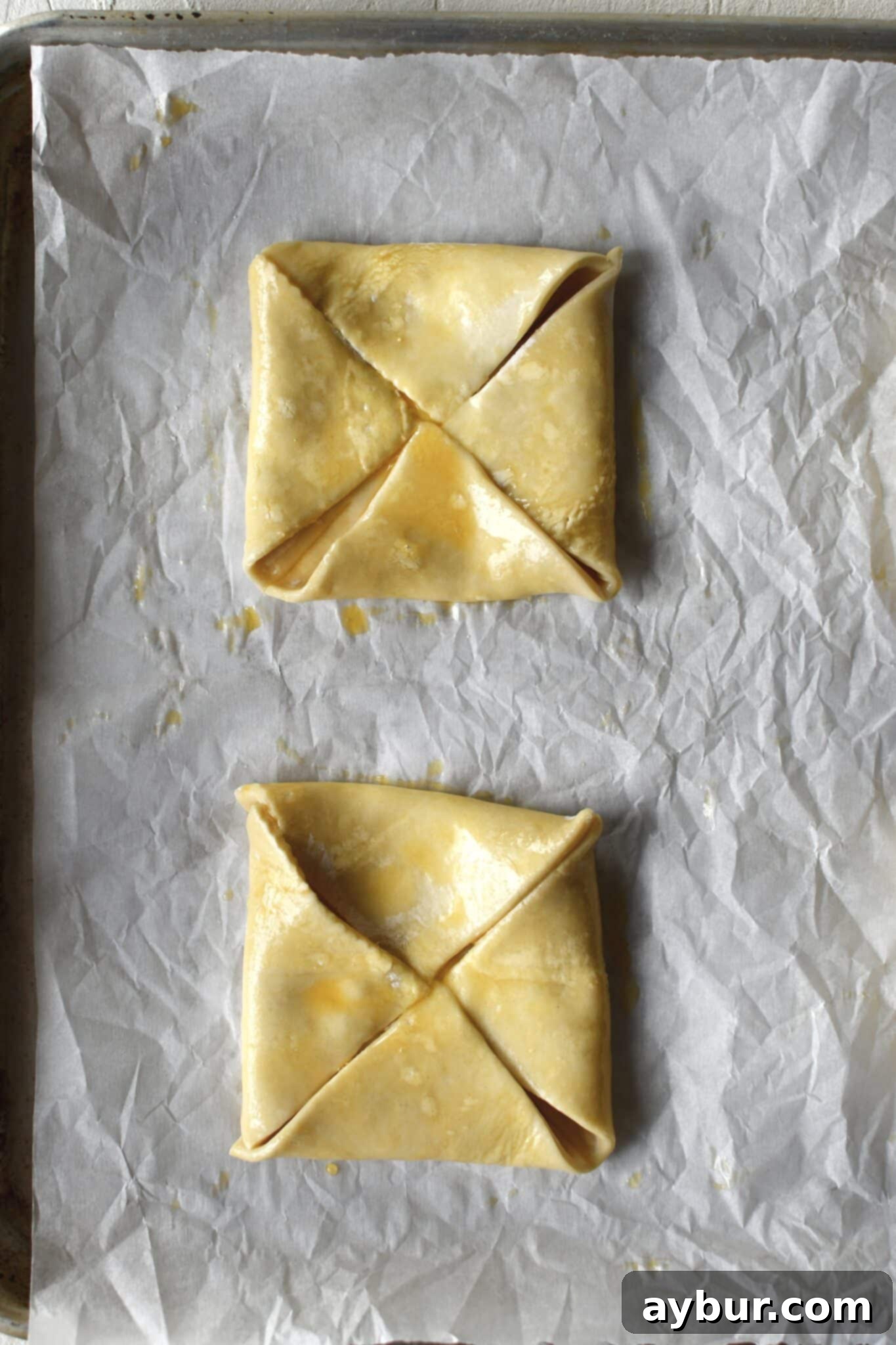 Neatly folded puff pastry squares, each corner brought to the center, lightly brushed with egg wash, and ready for chilling to maintain their shape.