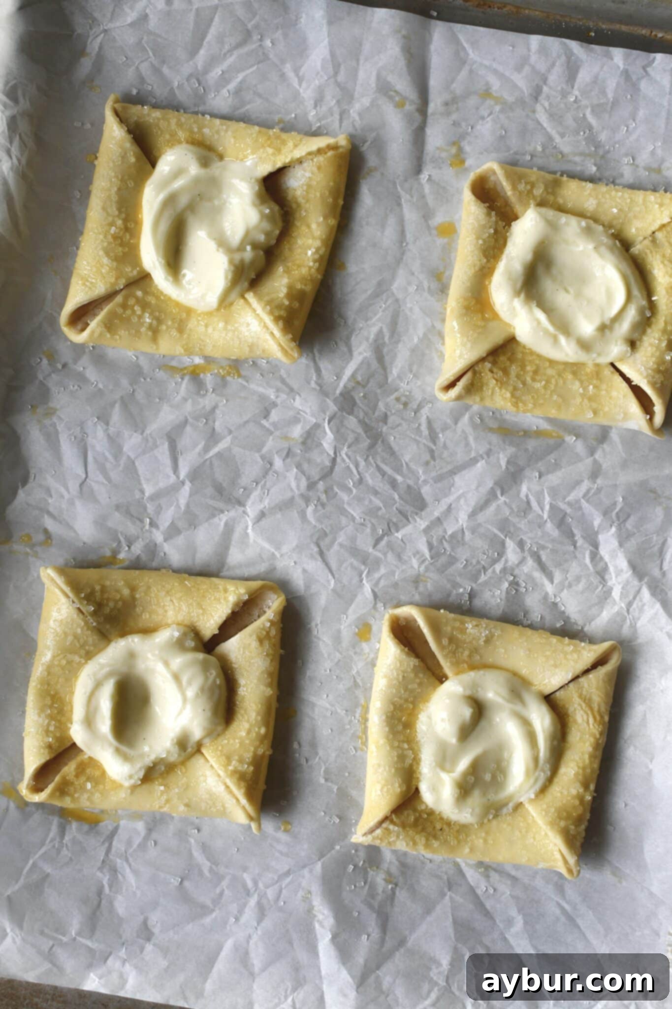 Chilled danishes, perfectly formed with a dollop of creamy cheese filling in the center of each, sprinkled with sanding sugar, and ready for baking.