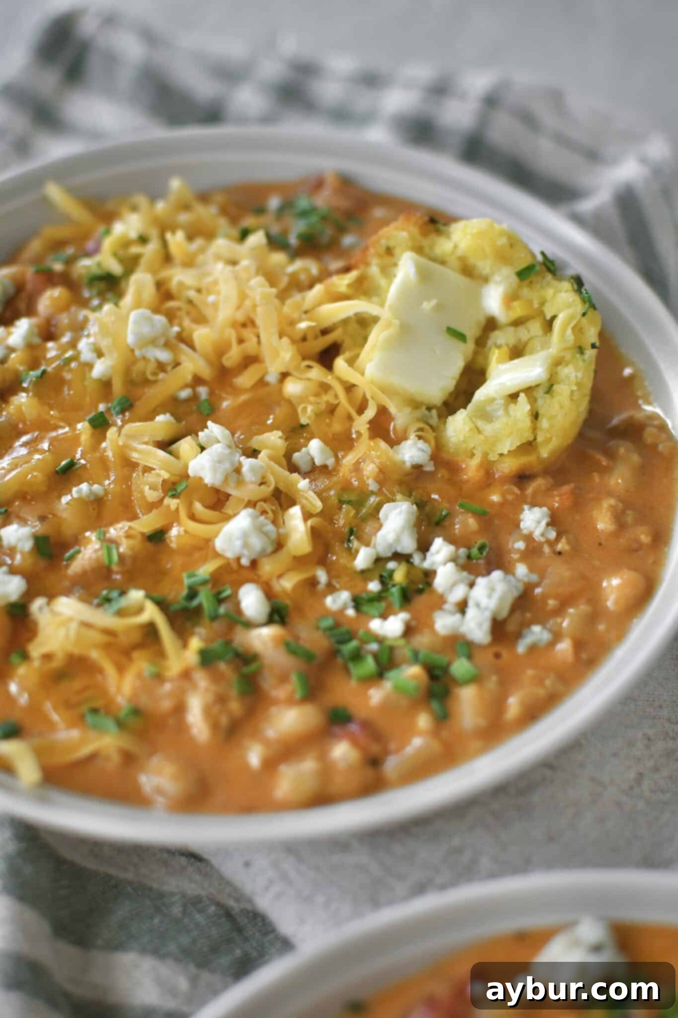 A steaming bowl of Buffalo Chicken Chili, generously topped with fresh chives, crumbled blue cheese, and shredded cheddar cheese. A piece of golden cornbread muffin is artfully dunked into the rich chili.