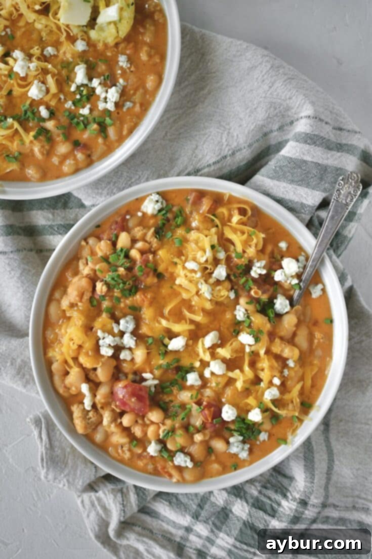 A beautifully presented bowl of Buffalo Chicken Chili, garnished with fresh chives, blue cheese crumbles, and shredded cheddar cheese. A piece of cornbread muffin is dipped into the rich chili.