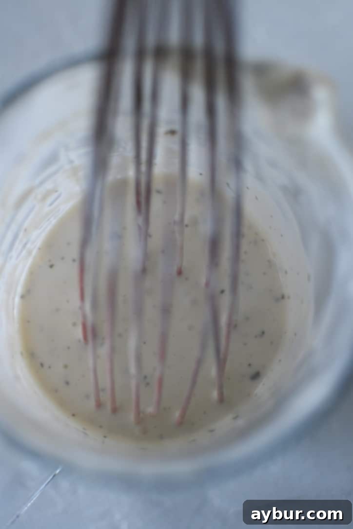 A whisk stirring creamy ranch dressing in a glass measuring cup.