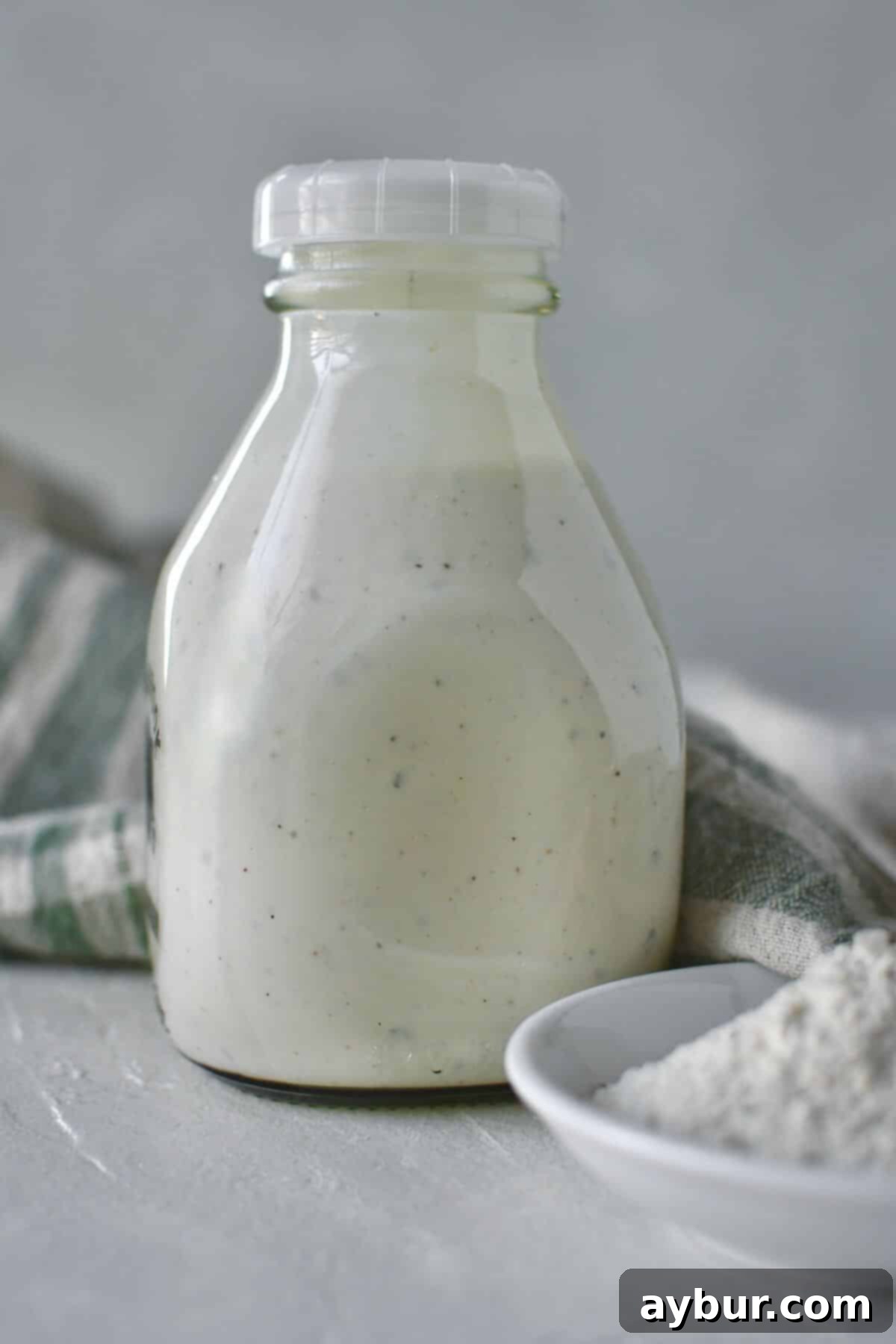 Freshly made ranch dressing in a clear glass bottle with a label, surrounded by fresh vegetables like carrots and celery sticks, ready for dipping.