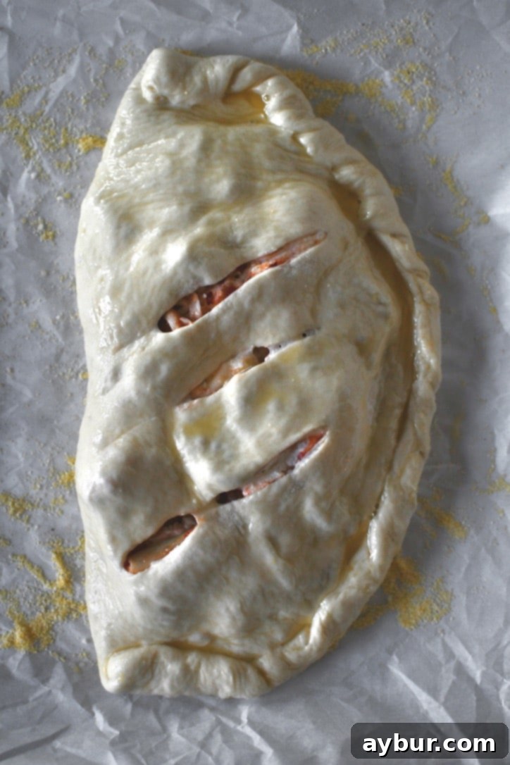 Folded calzone dough with crimped edges and three slits to release steam, ready for baking after being brushed with egg wash.