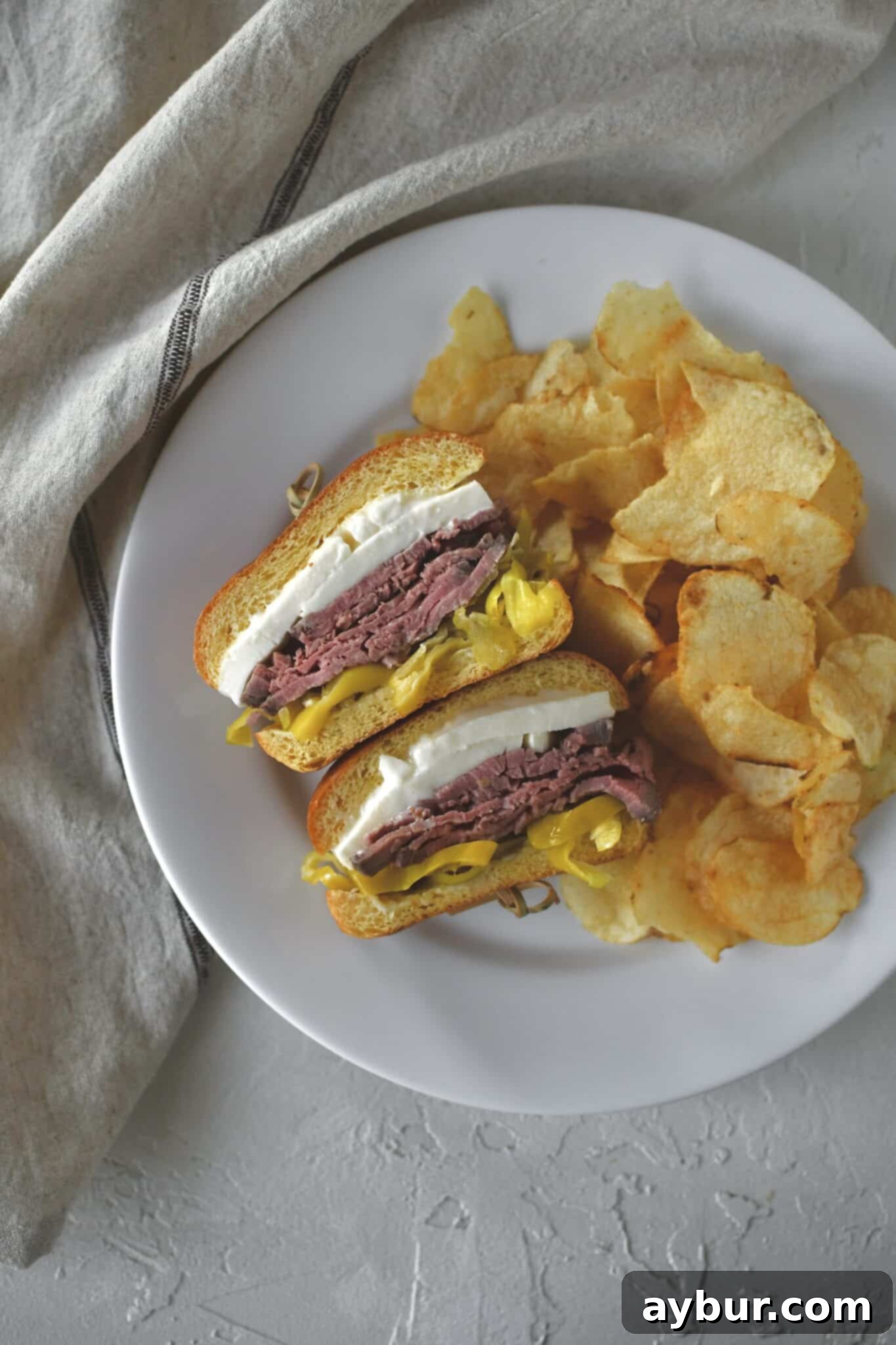 A beautifully assembled roast beef sandwich, sliced in half to reveal its layers, served on a plate with crispy potato chips, ready to be enjoyed.